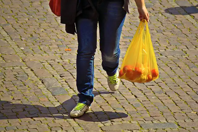 Man erkennt einen Menschen in Straßenkleidung, der eine Plastiktüte mit Einkäufen trägt. Gezeigt werden nur die Beine.